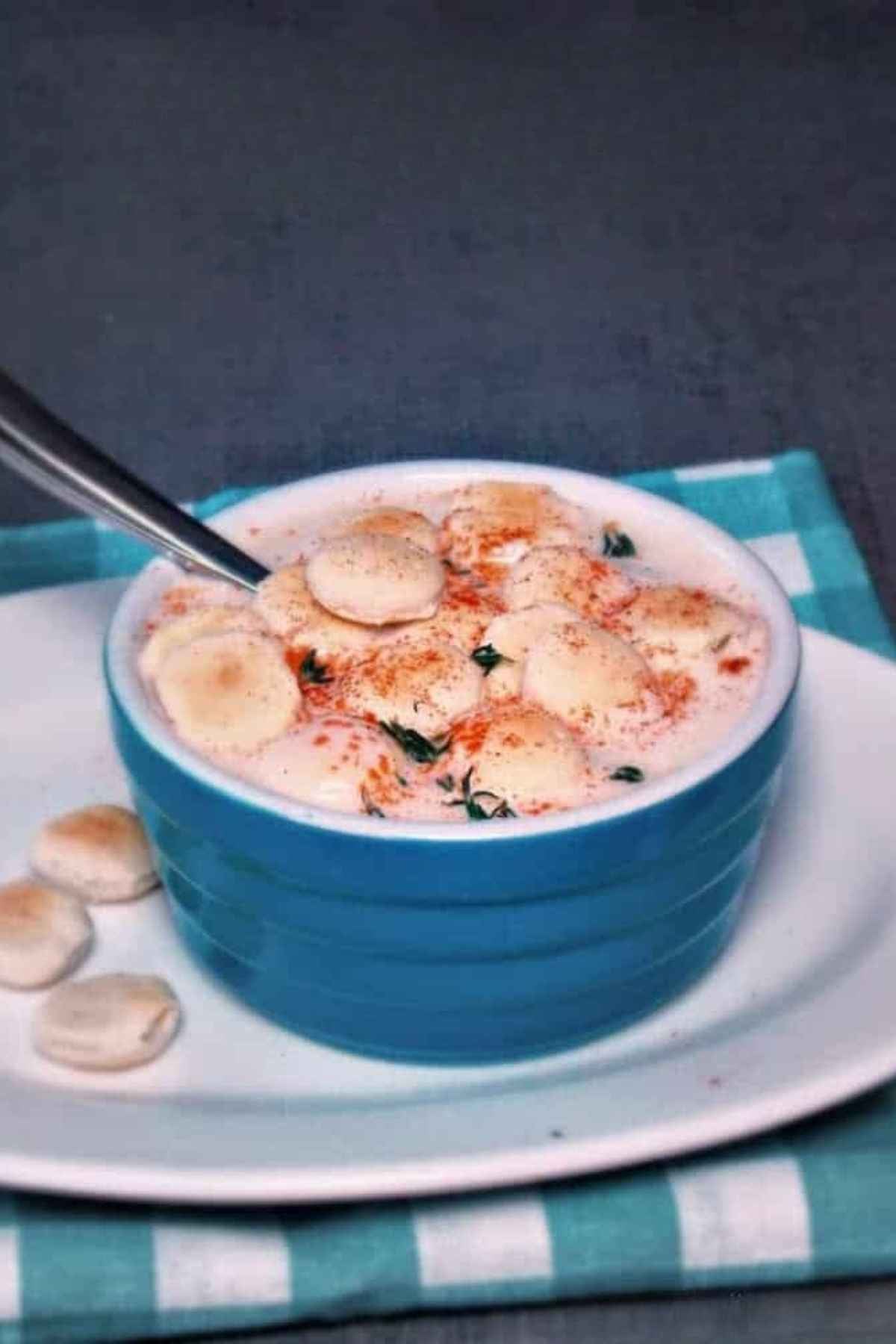 A blue ceramic bowl filled with creamy slow cooker clam chowder topped with several oyster crackers and a sprinkle of paprika, served with a spoon on a blue checkered napkin.