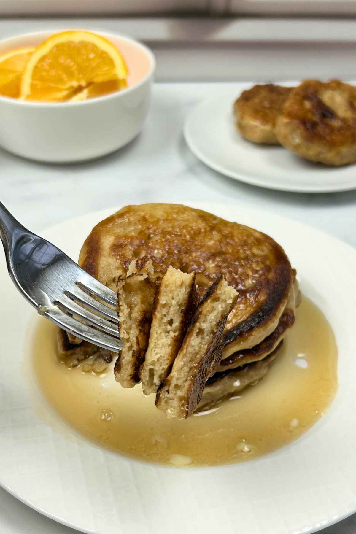 A close up shot of a stack of small, golden-brown buttermilk pancake bites on a white plate, with a fork lifting a few pieces to show the fluffy interior, all drizzled with maple syrup.