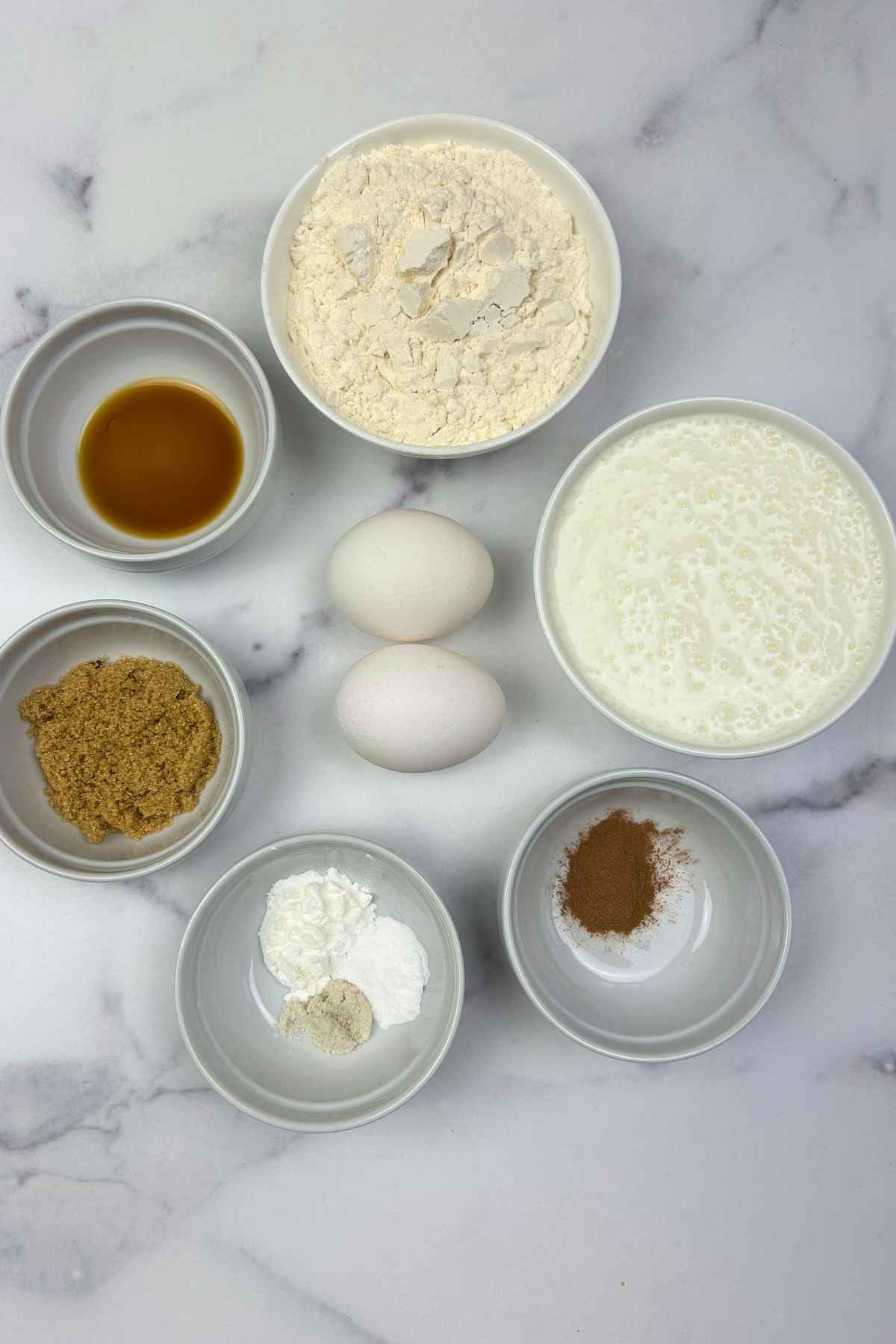 An assortment of baking essentials including flour, eggs, buttermilk, and spices in prep bowls on a marble background.