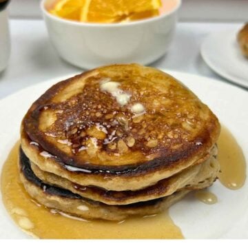 A tall stack of three thick, fluffy griddle cakes topped with a melting pat of butter and glistening amber syrup, served on a white plate with a side of fresh orange slices.