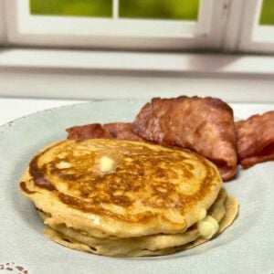 Close-up of aerated, toasted breakfast cakes with a square melting butter and side of wavy, browned bacon strips on a patterned plate.