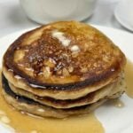 A tall stack of three thick, fluffy griddle cakes topped with a melting pat of butter and glistening amber syrup, served on a white plate with a side of fresh orange slices.