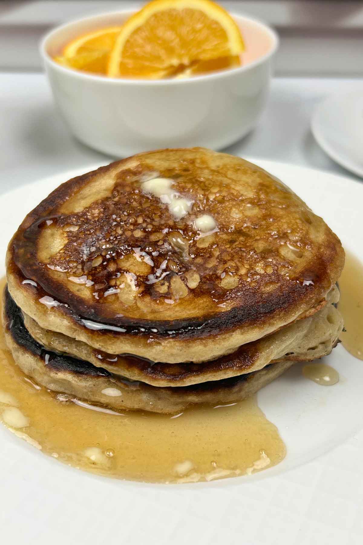 A tall stack of three thick, fluffy griddle cakes topped with a melting pat of butter and glistening amber syrup, served on a white plate with a side of fresh orange slices.