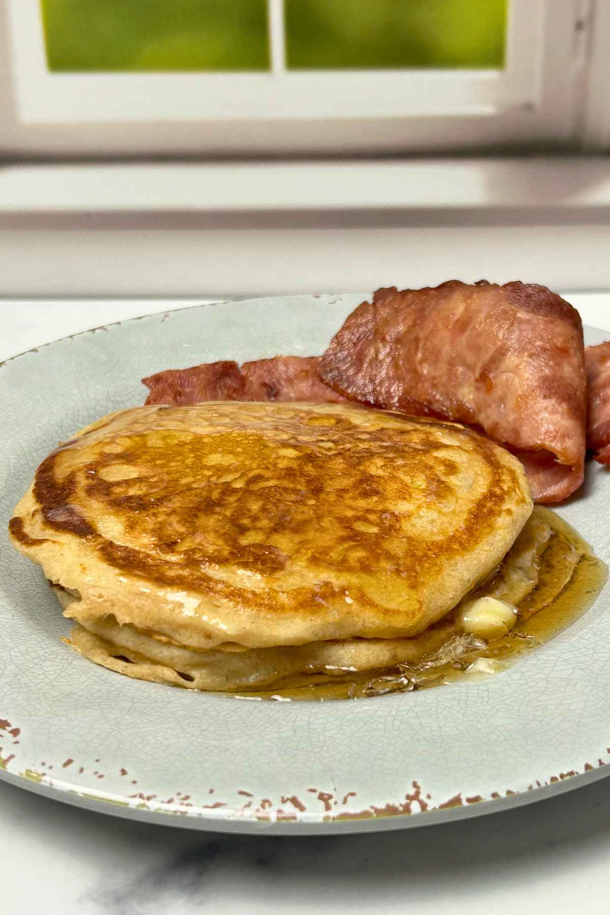 Close-up of aerated, buttermilk pancakes with a square of melting butter and side of wavy, browned bacon strips on a patterned plate.