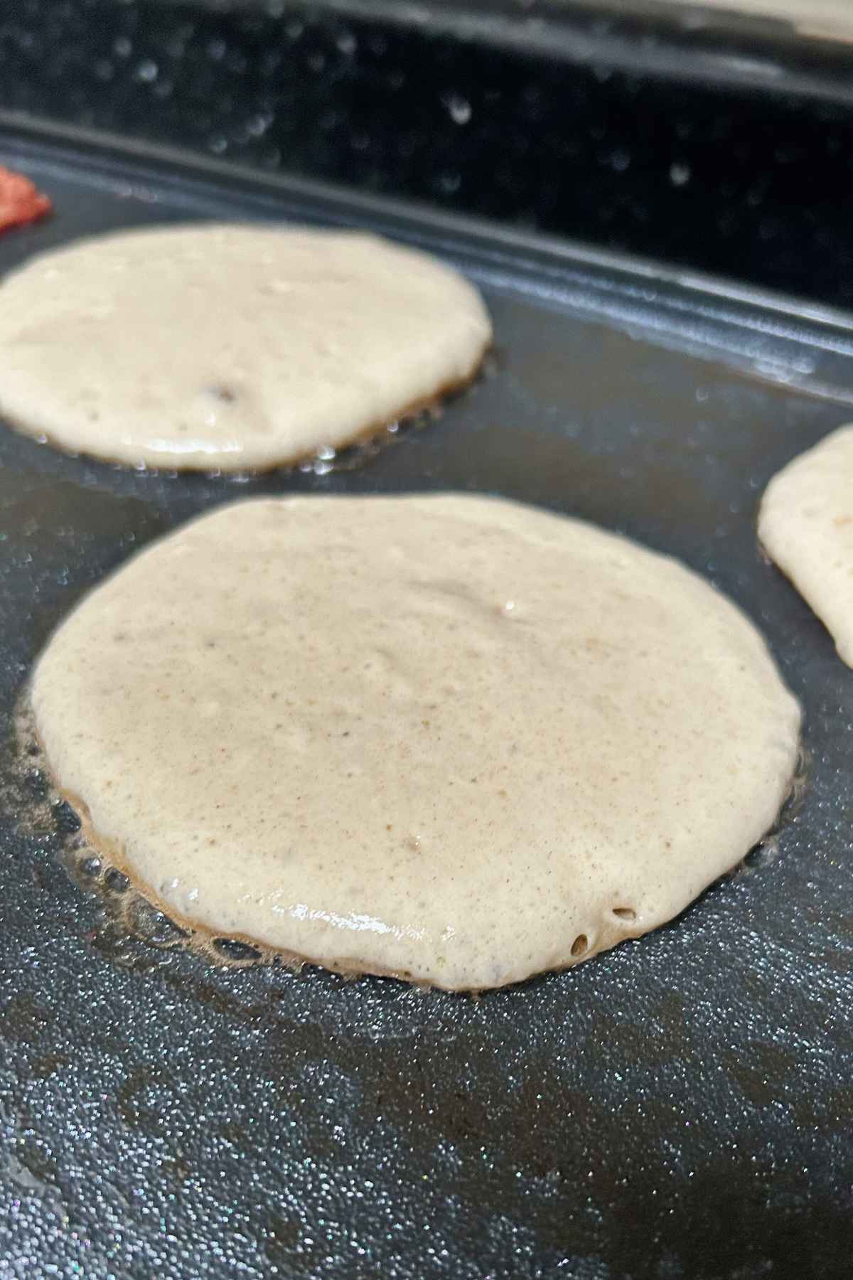 Close up of buttermilk pancake batter on a flat-top grill, featuring a smooth, cream colored surface with visible leavening bubbles.