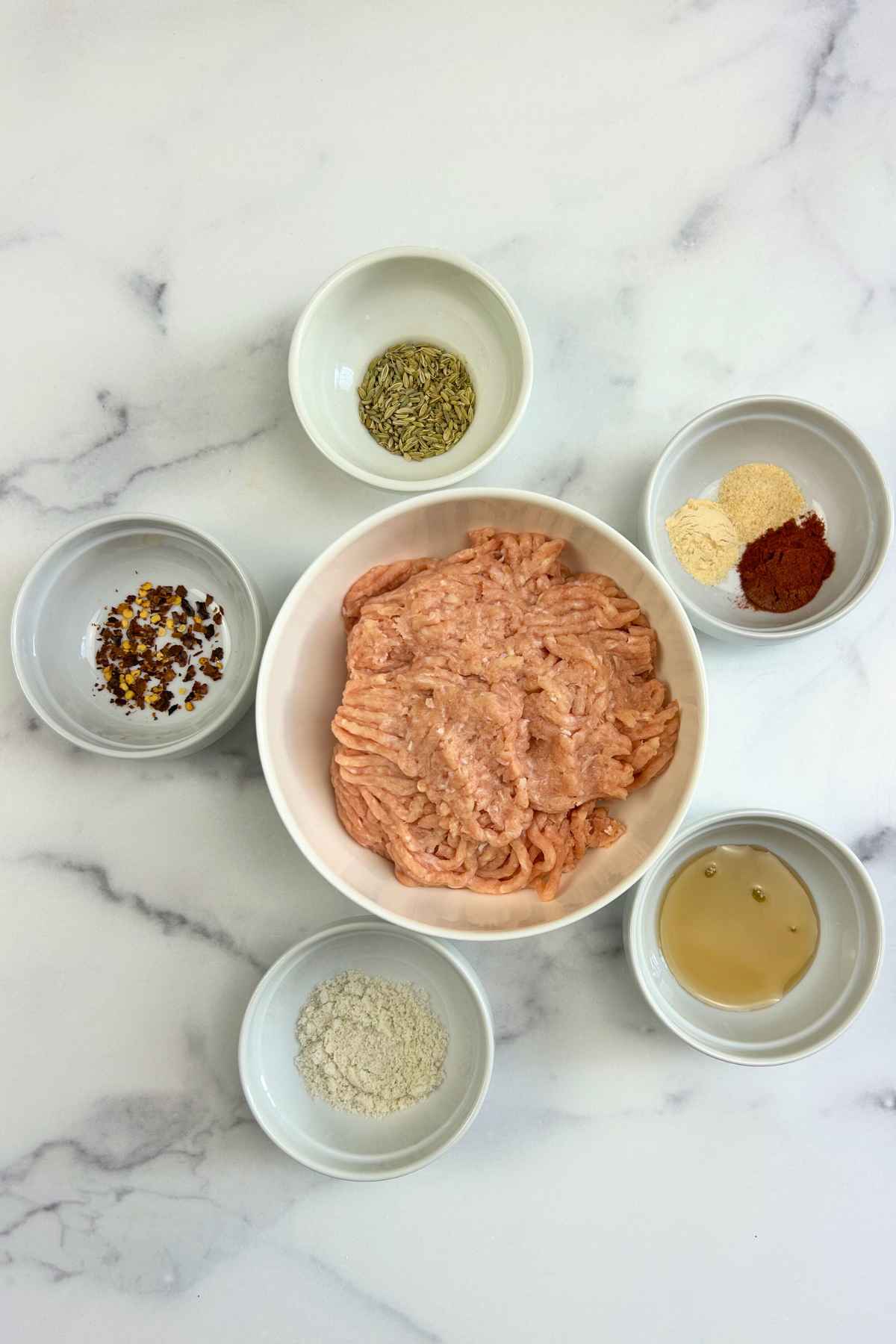 Raw ground poultry in a white bowl surrounded by small dishes of fennel seeds, red pepper flakes, garlic powder, smoked paprika, and maple syrup.