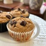 A close-up of high-protein peanut butter banana chocolate chips muffins in paper liners, topped with melted semi sweet chocolate chips on a white plate.