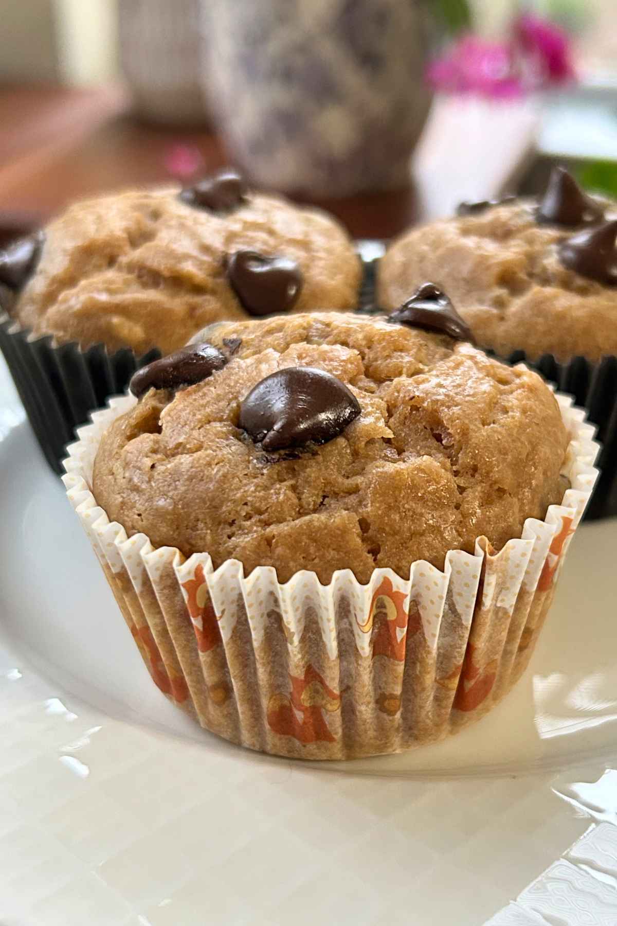 A close up of three soft muffins in patterned paper l iners, topped with large melted chocolate chips on a white ceramic plate.
