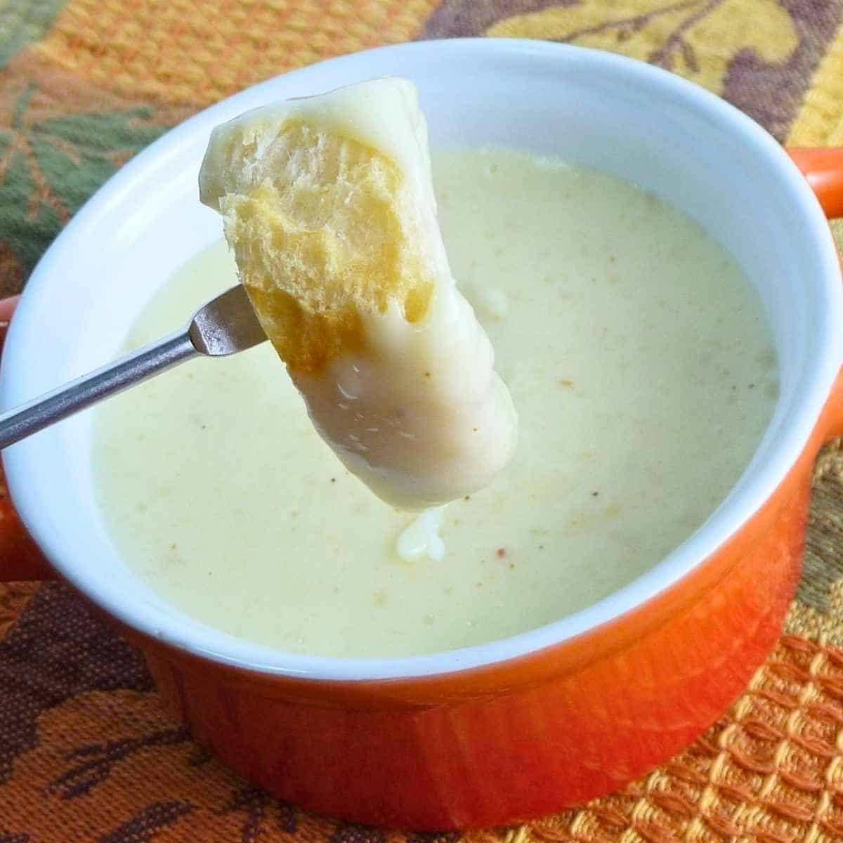 A crusty bread cube dipped into a creamy white cheese fondue in an orange ceramic pot.