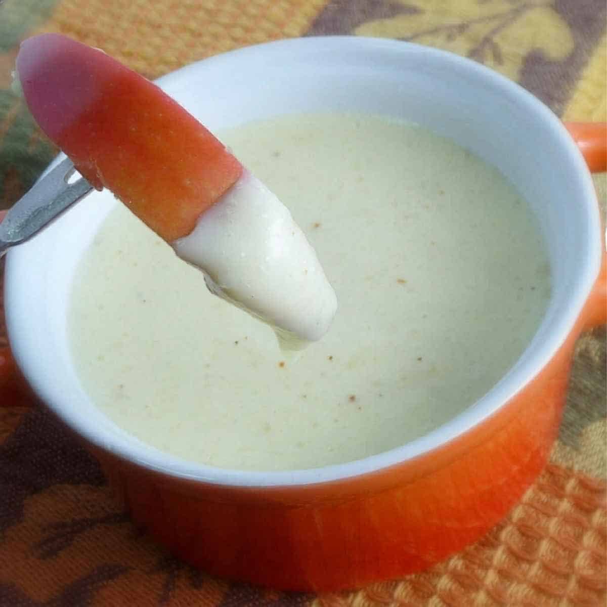 A crisp apple wedge being dipped into a bowl of smooth, melted cheese fondue dip.