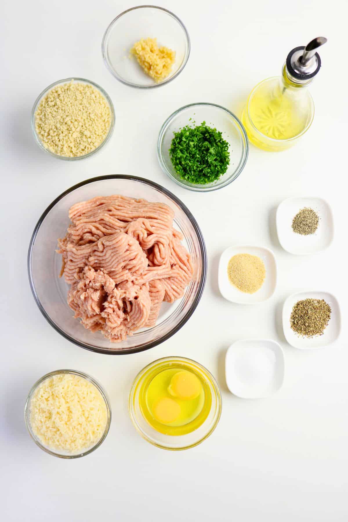 A flat lay of measured ingredients for a ground chicken recipe, including meat, eggs, herbs, and cheese in clear glass prep bowls.