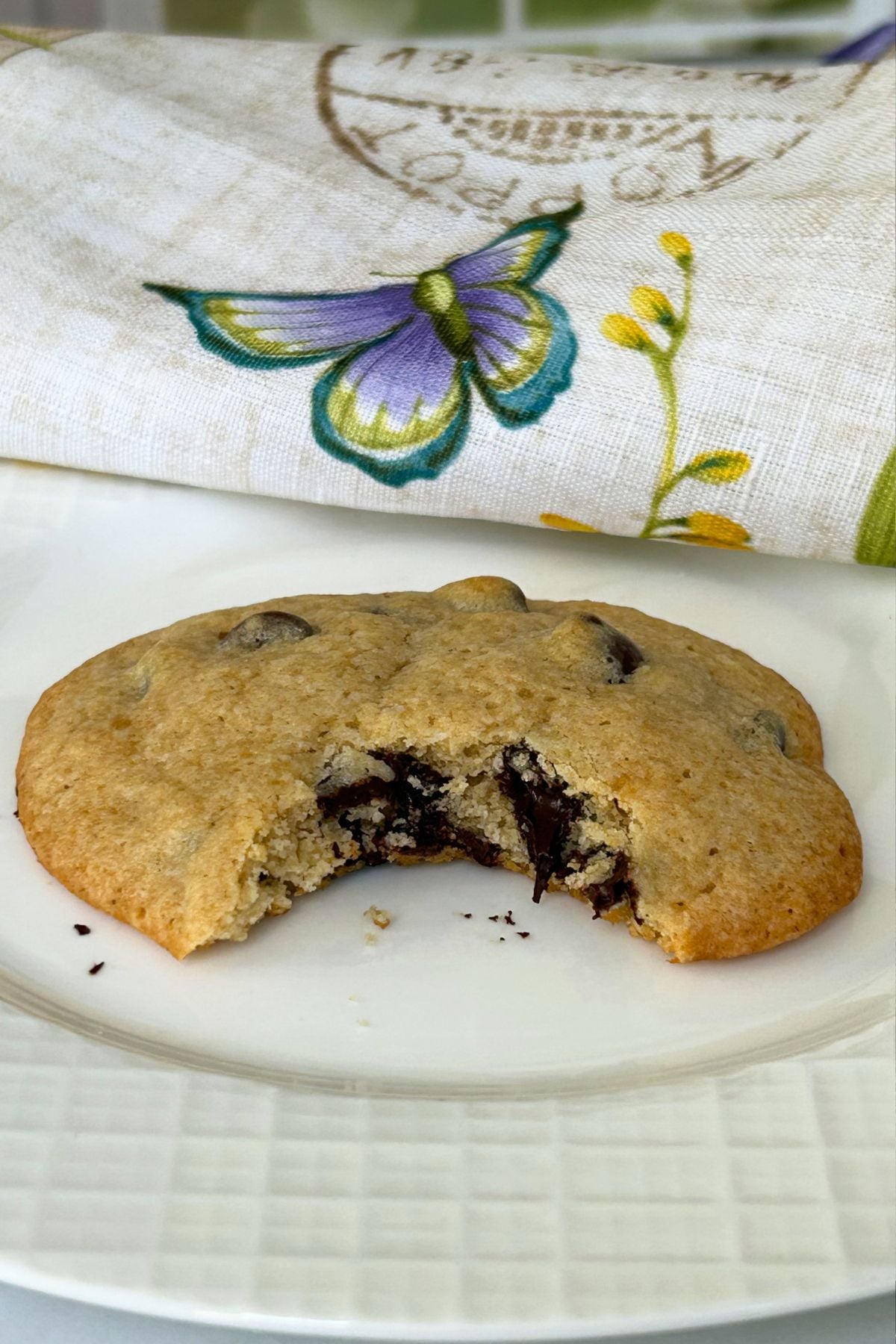 A single cookie on a white plate with a bite taken out of the cookie, revealing the texture and melted chocolate chips inside.