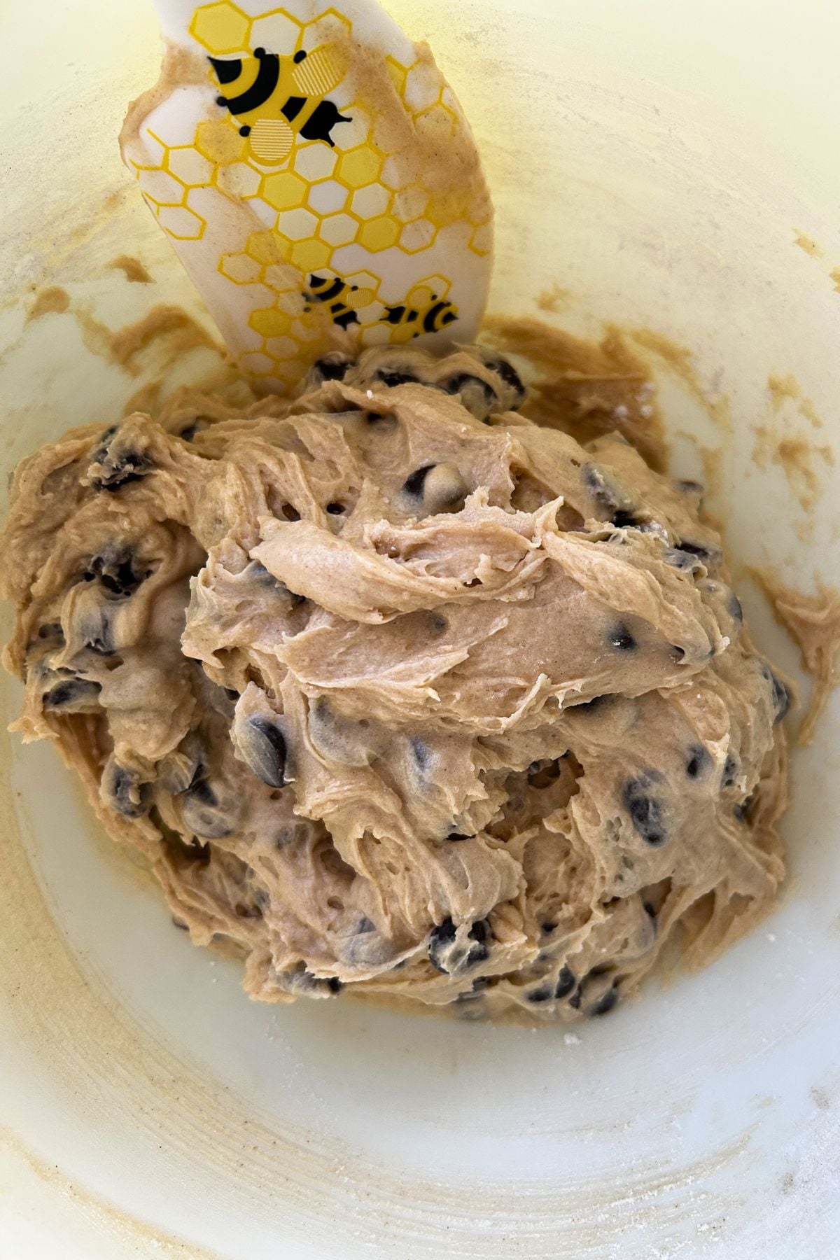 A bowl of thick cookie dough filled with chocolate chips with a white spatula featuring a yellow honeycomb and black bumblebee design rests in the bowl.