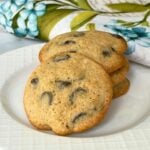 A stack of thick, soft chocolate chip cookies served on a white plate with a floral and butterfly embroidered napkin in the background.