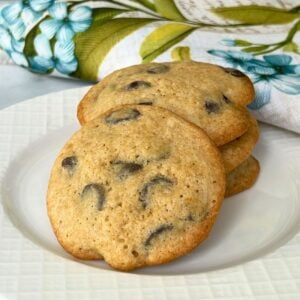 A stack of thick, soft chocolate chip cookies served on a white plate with a floral and butterfly embroidered napkin in the background.