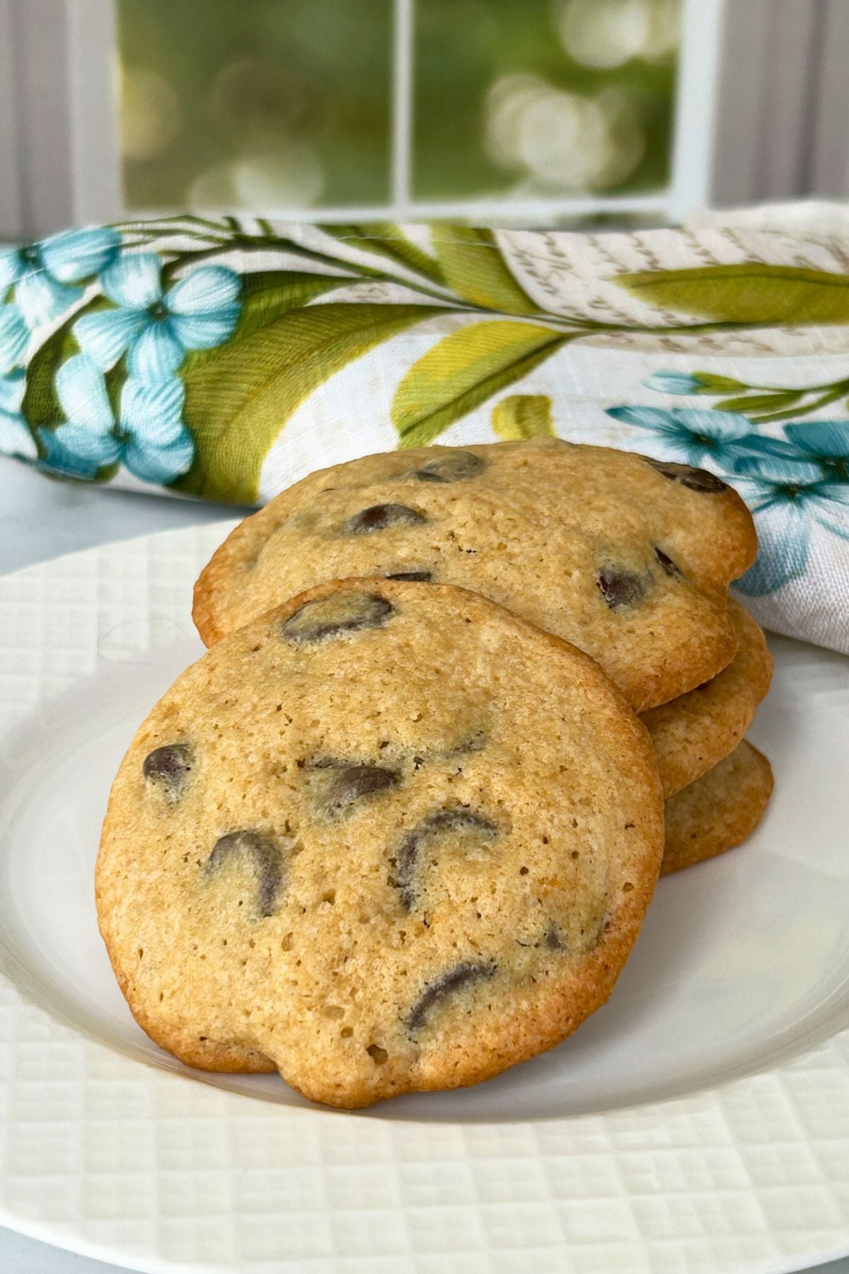 A stack of thick, soft chocolate chip cookies served on a white plate with a floral and butterfly embroidered napkin in the background.