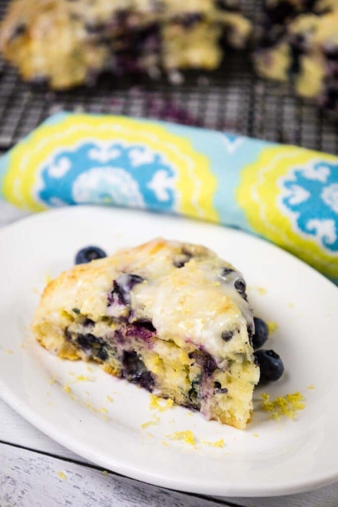 A glazed blueberry scone with lemon zest on a white plate, positioned next to a blue and yellow patterned napkin.