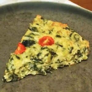 A slice of kohlrabi greens quiche with red cherry tomato pieces on a dark plate.