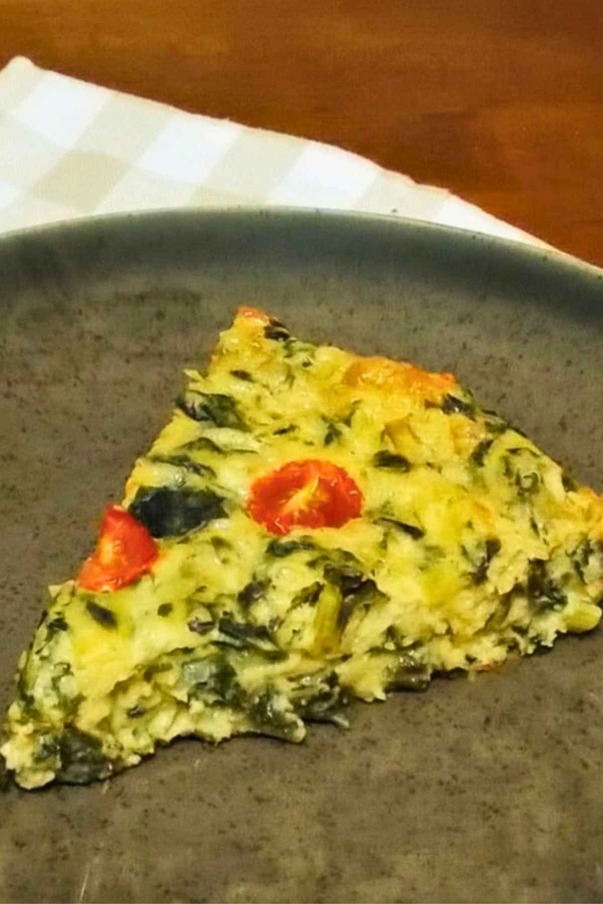 A slice of kohlrabi greens quiche with red cherry tomato pieces on a dark plate.