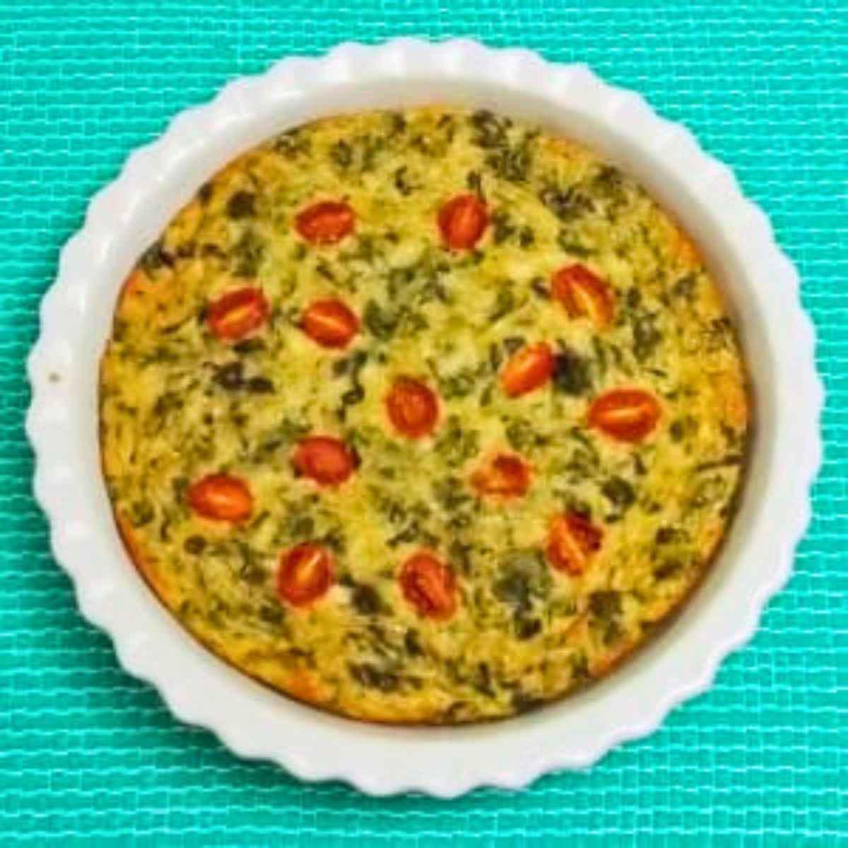 Overhead view of a quiche in a round white ceramic pan, dotted with halved cherry tomatoes, set on a teal textured surface.