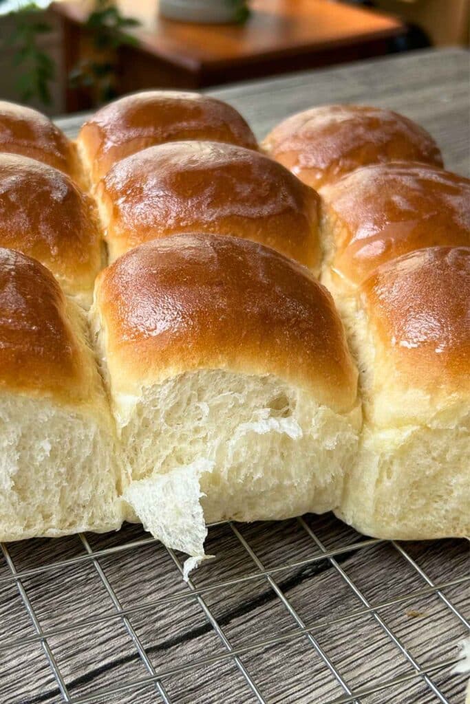 A close-up of golden brown, fluffy sweet dinner rolls cooling on a wire rack, showing a soft, flaked texture where one roll has been pulled a part.