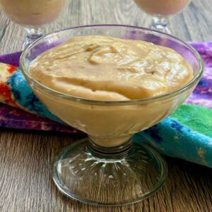 Clear glass footed dish filled with creamy homemade butterscotch pudding on a colorful tea towel.