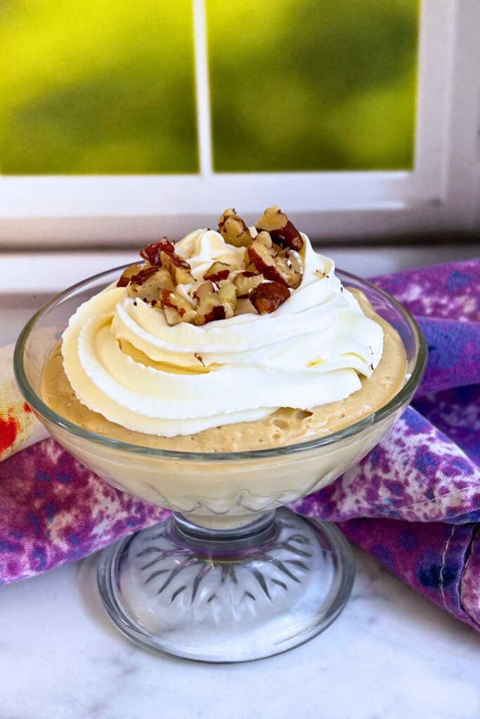 Clear glass compote dish filled with butterscotch pudding, topped with whipped cream swirls and chopped pecans.