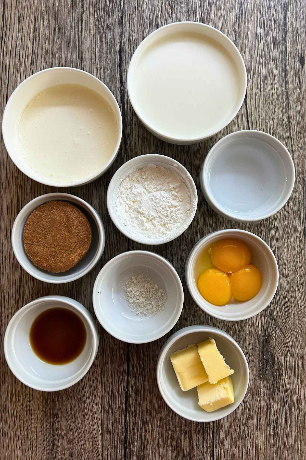 Small white prep bowls filled with brown sugar, egg yolks, butter, cornstarch, milk, and vanilla arranged on a wooden surface.