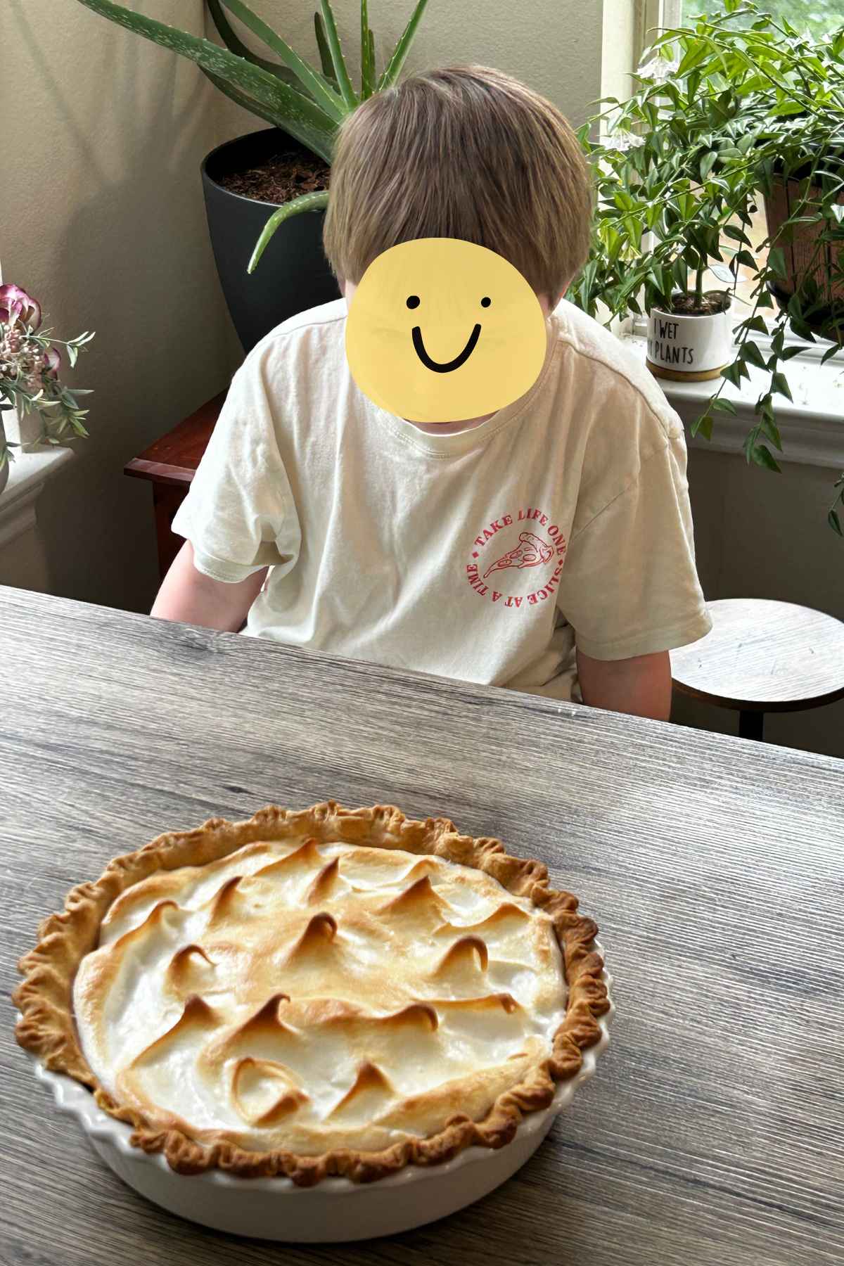 A young child intensely staring at a butterscotch meringue pie on a wooden table.