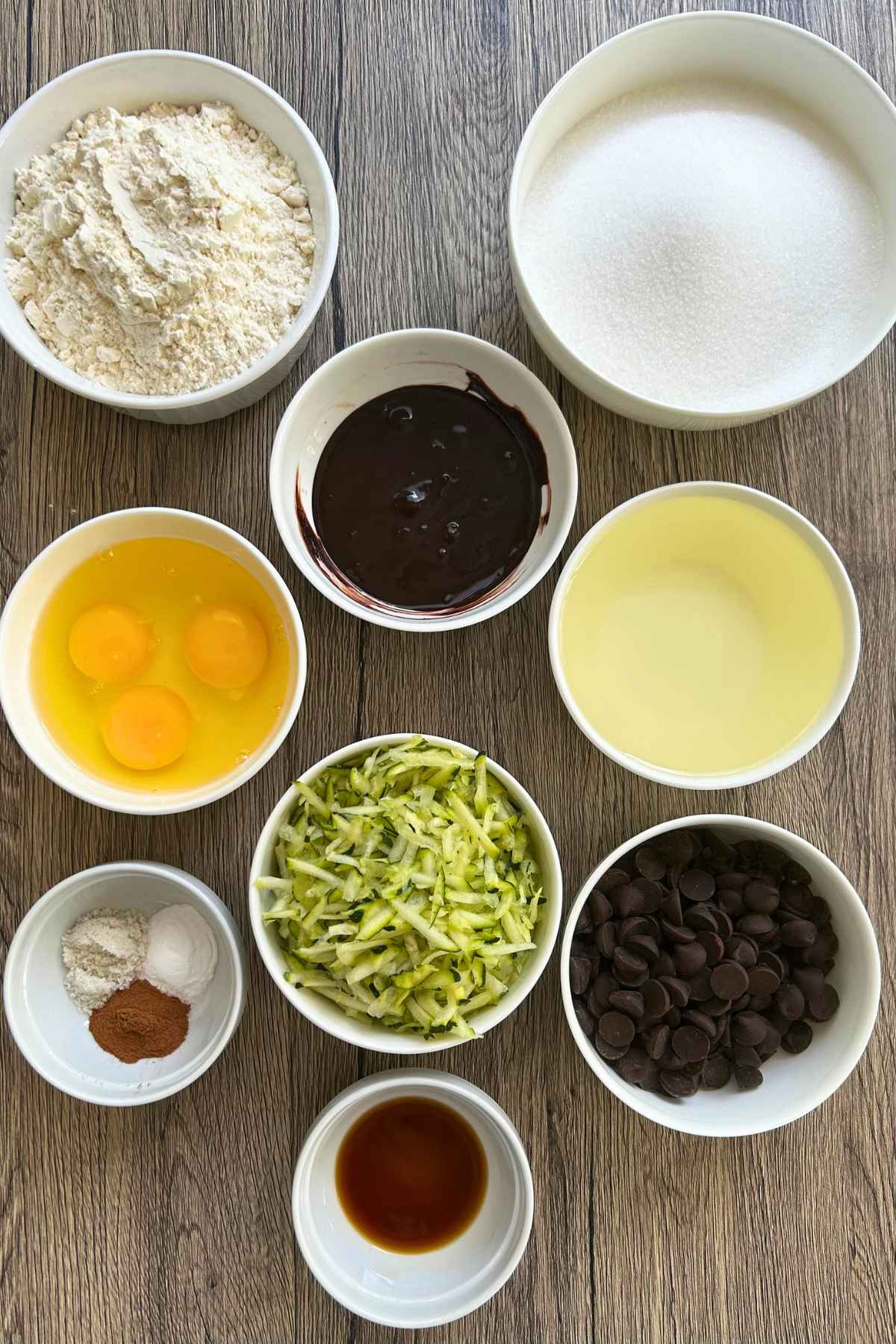 Overhead view of small white bowls containing flour, sugar, oil, eggs, grated zucchini, chocolate chips, and spices arranged on a dark surface.