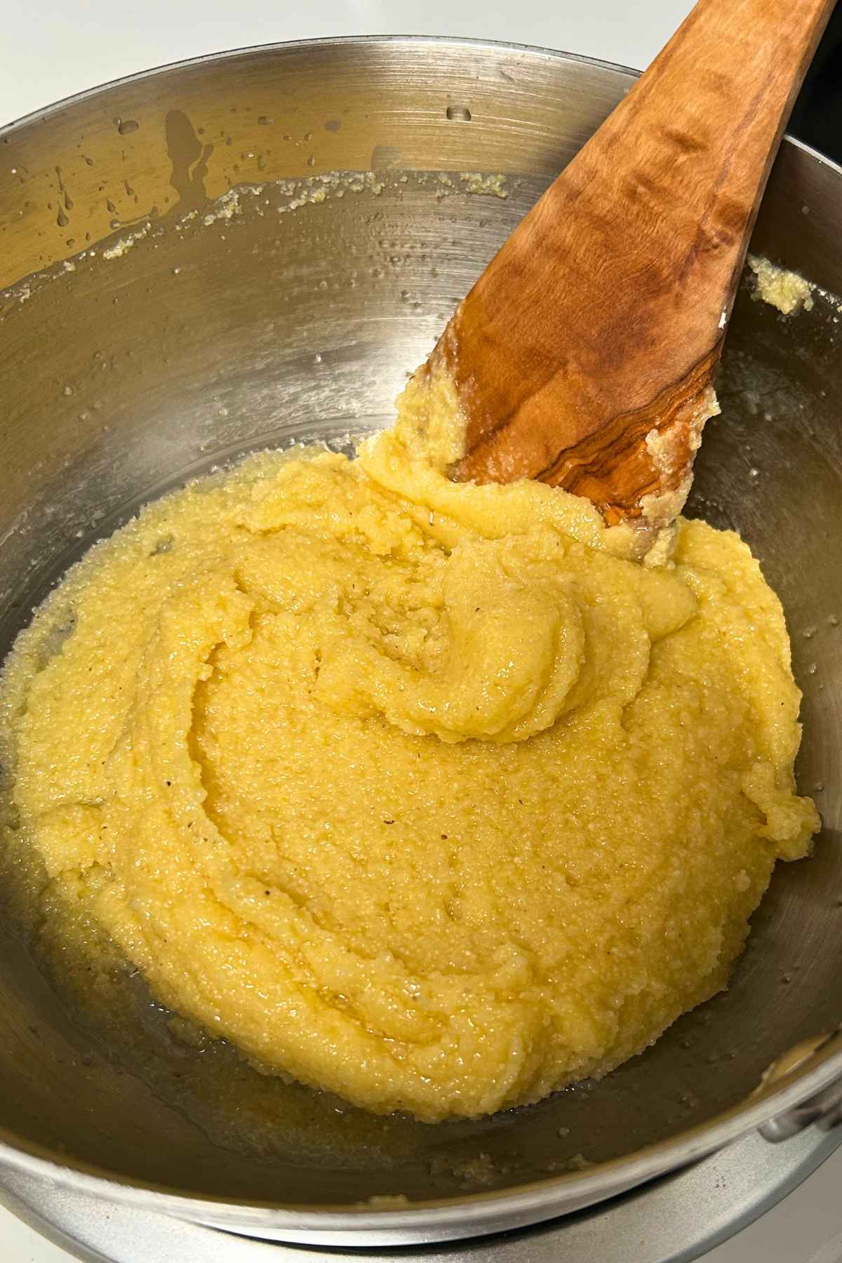 A wooden spatula stirring a thick, vibrant yellow cornmeal dough mixture inside a stainless steel mixing bowl.