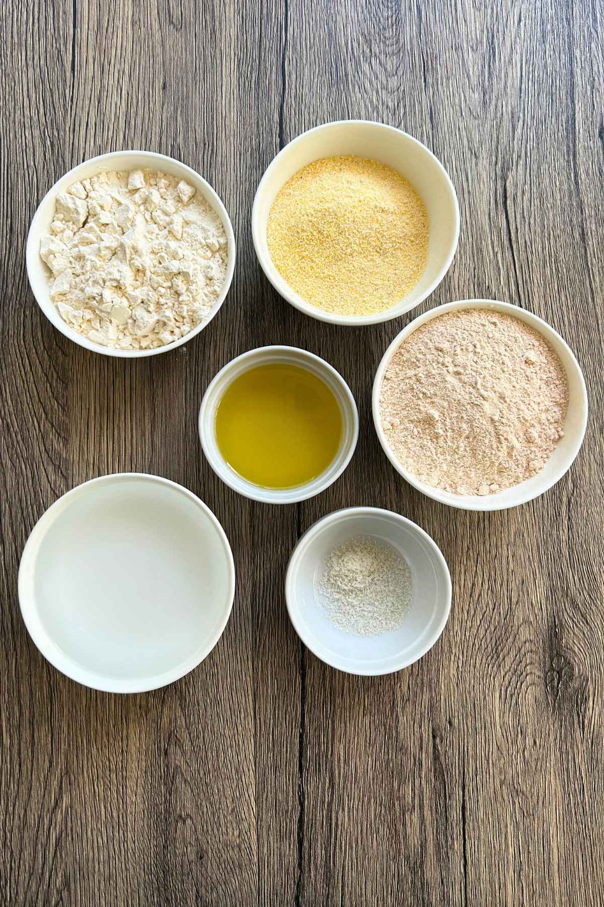 Six small white ceramic bowls arranged on a wooden surface, containing ingredients like flour, cornmeal, olive oil and salt.