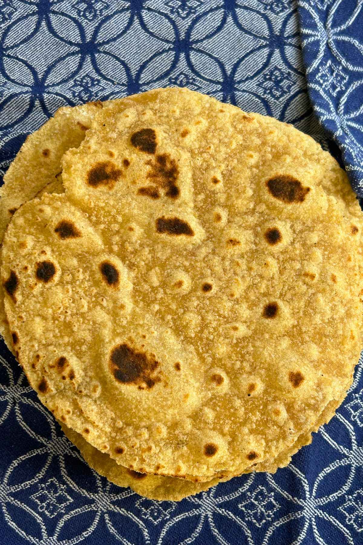 Overhead image of a stack of freshly cooked golden brown corn and flour tortillas with dark charred spots, resting on a blue and white patterned cloth napkin.