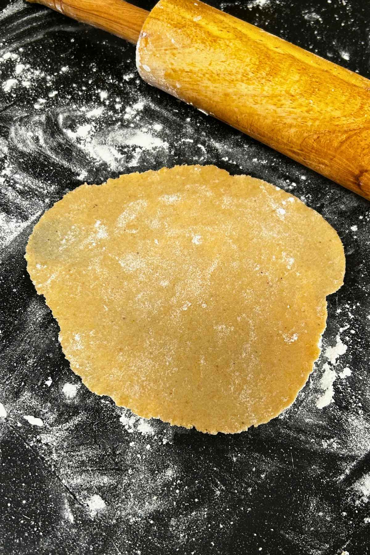 A rolled out circular piece of uncooked tortilla dough resting on a dark, flour dusted countertop next to a wooden rolling pin.