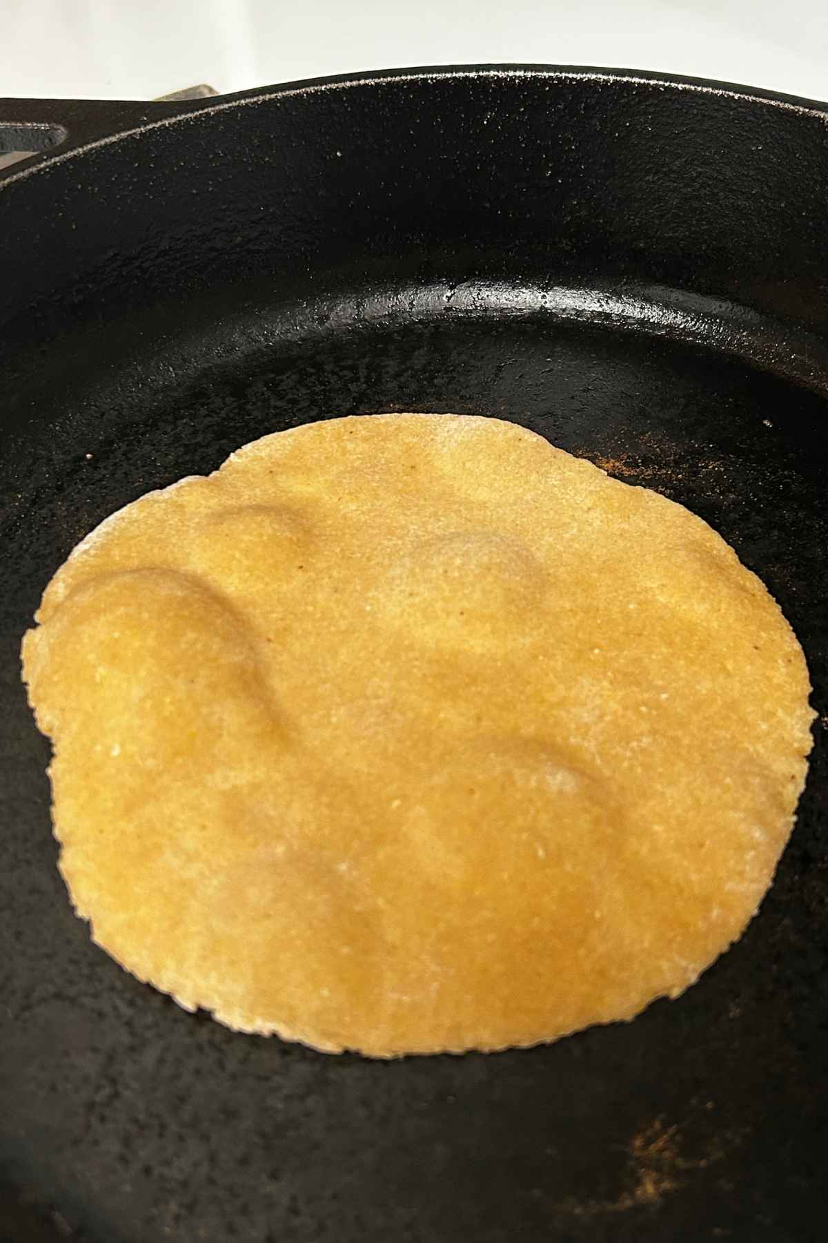 A homemade corn and flour tortilla frying and puffing up inside a hot, seasoned black cast iron skillet.