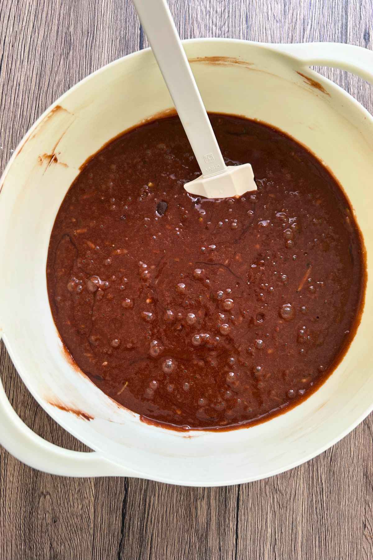 Rich, dark chocolate batter with visible shreds of zucchini mixed in a large white bowl with a white spatula.