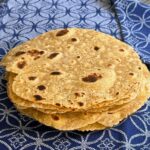 Overhead image of a stack of freshly cooked golden brown corn and flour tortillas with dark charred spots, resting on a blue and white patterned cloth napkin.