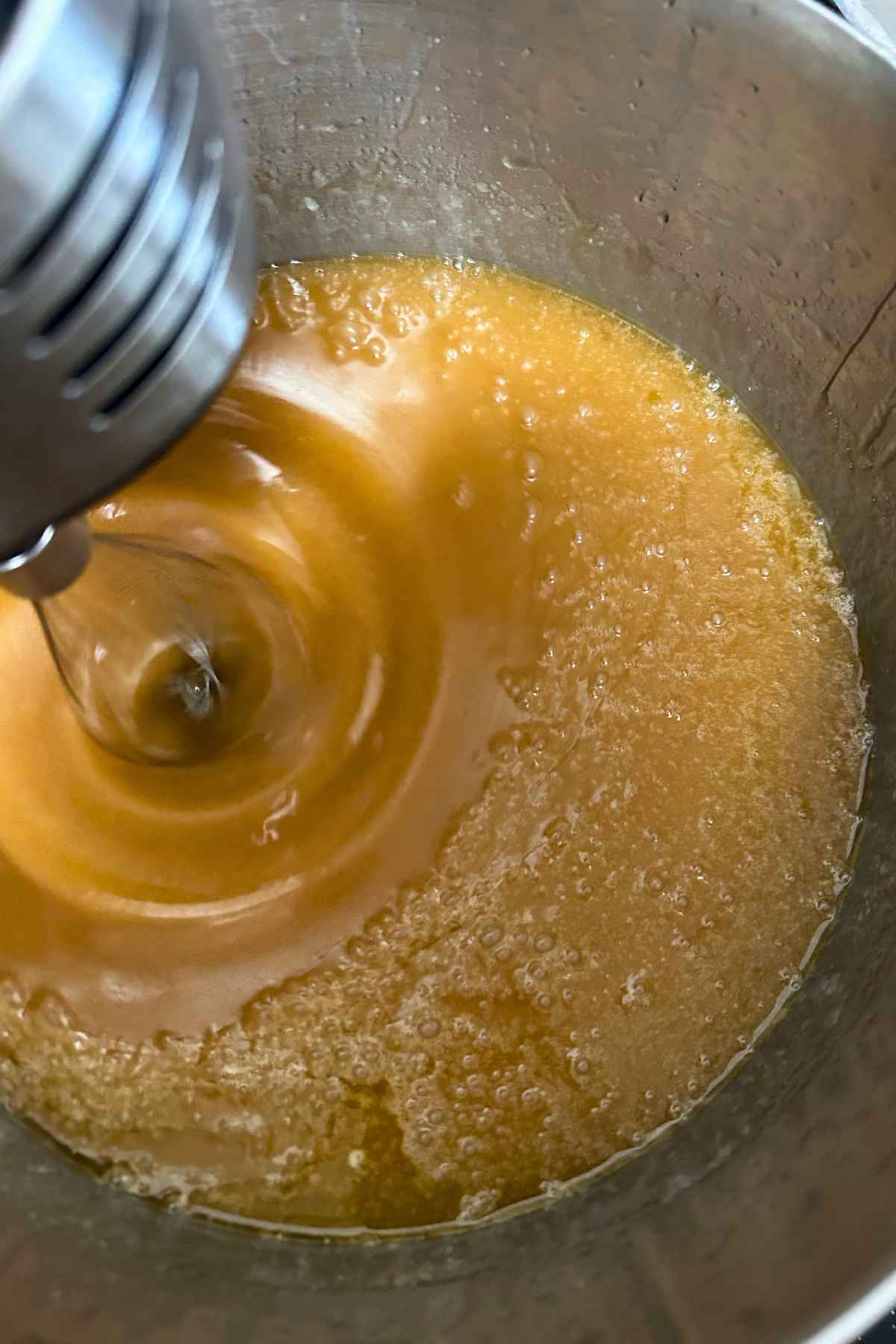 Electric hand mixer whisking warm boiled syrup and melted butter in a pot.
