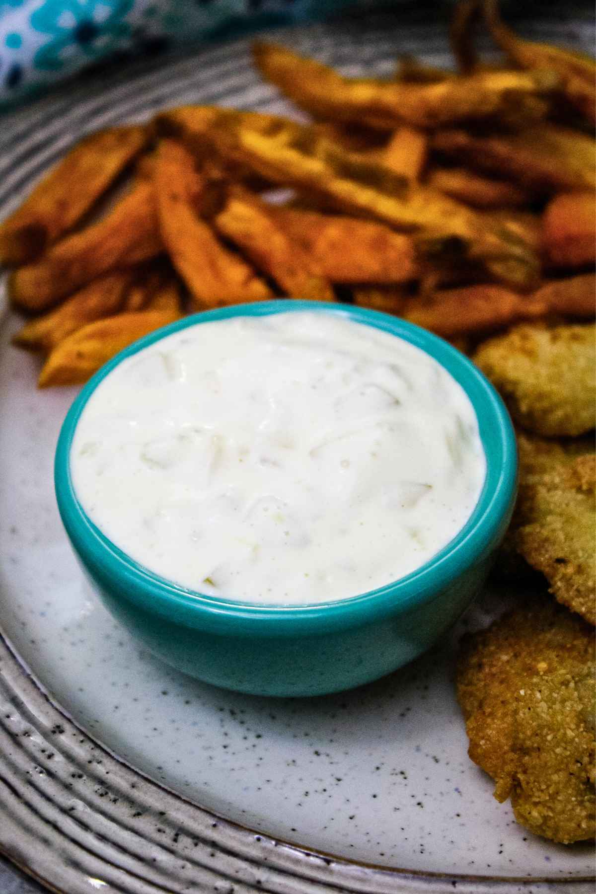 A small teal bowl filled with creamy homemade tartar sauce, served on a speckled plate next to crispy sweet potato fries and golden fish fillets.