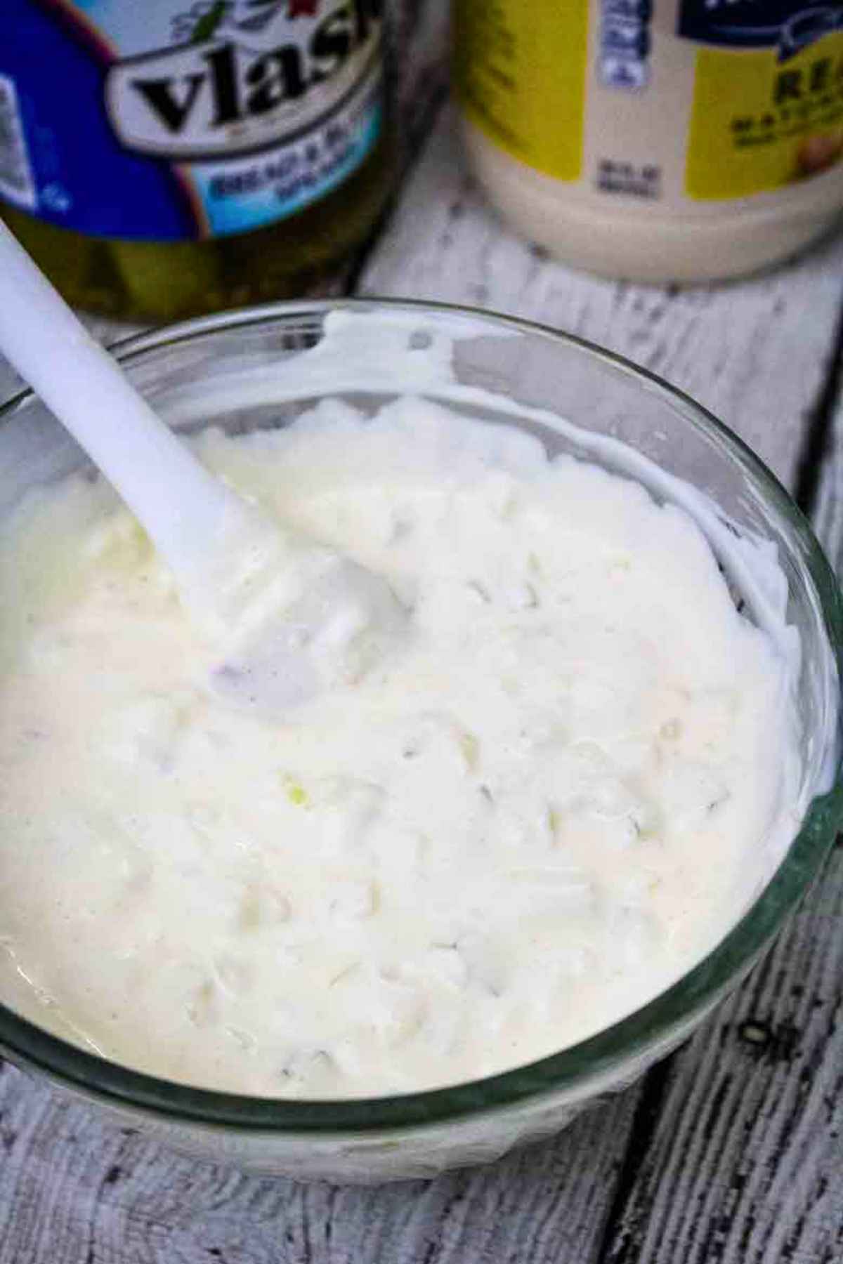 Freshly mixed homemad tartar sauce with finely chopped pickles and onions in a clear glass bowl with a white spatula.