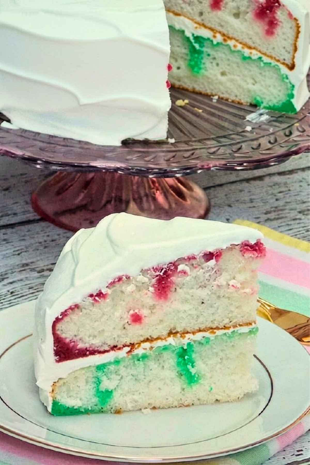 Slice of white jello poke cake with red and green fillings and white whipped cream frosting, served on a plate.