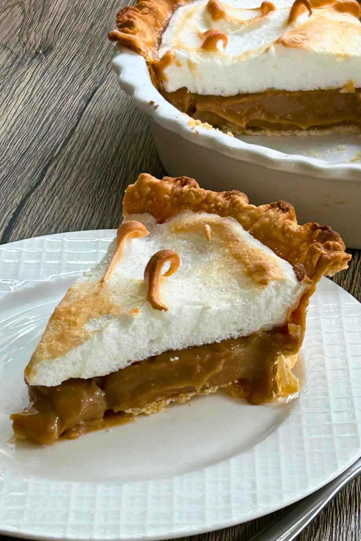 A slice of homemade butterscotch meringue pie on a white plate with the whole pie in the background.