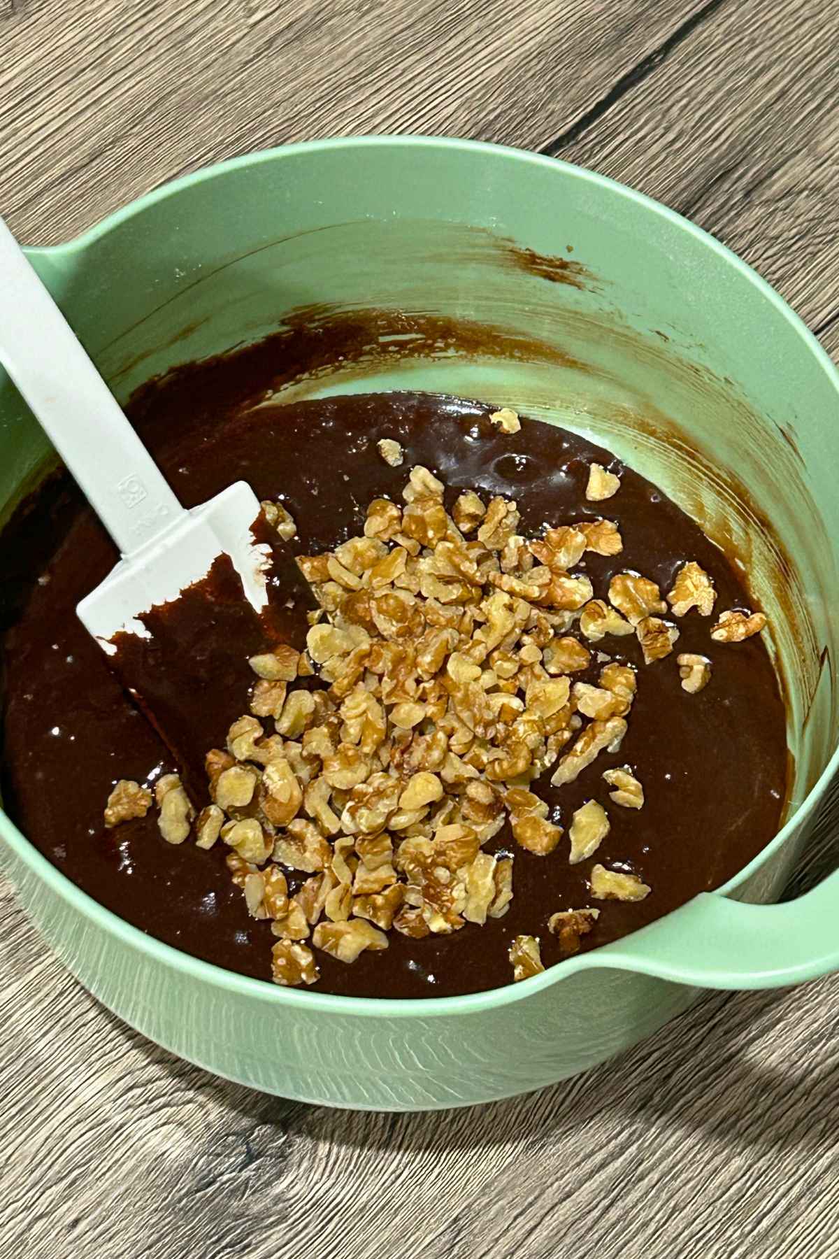A light green mixing bowl filled with dark chocolate batter topped with chopped nuts and a white spatula.