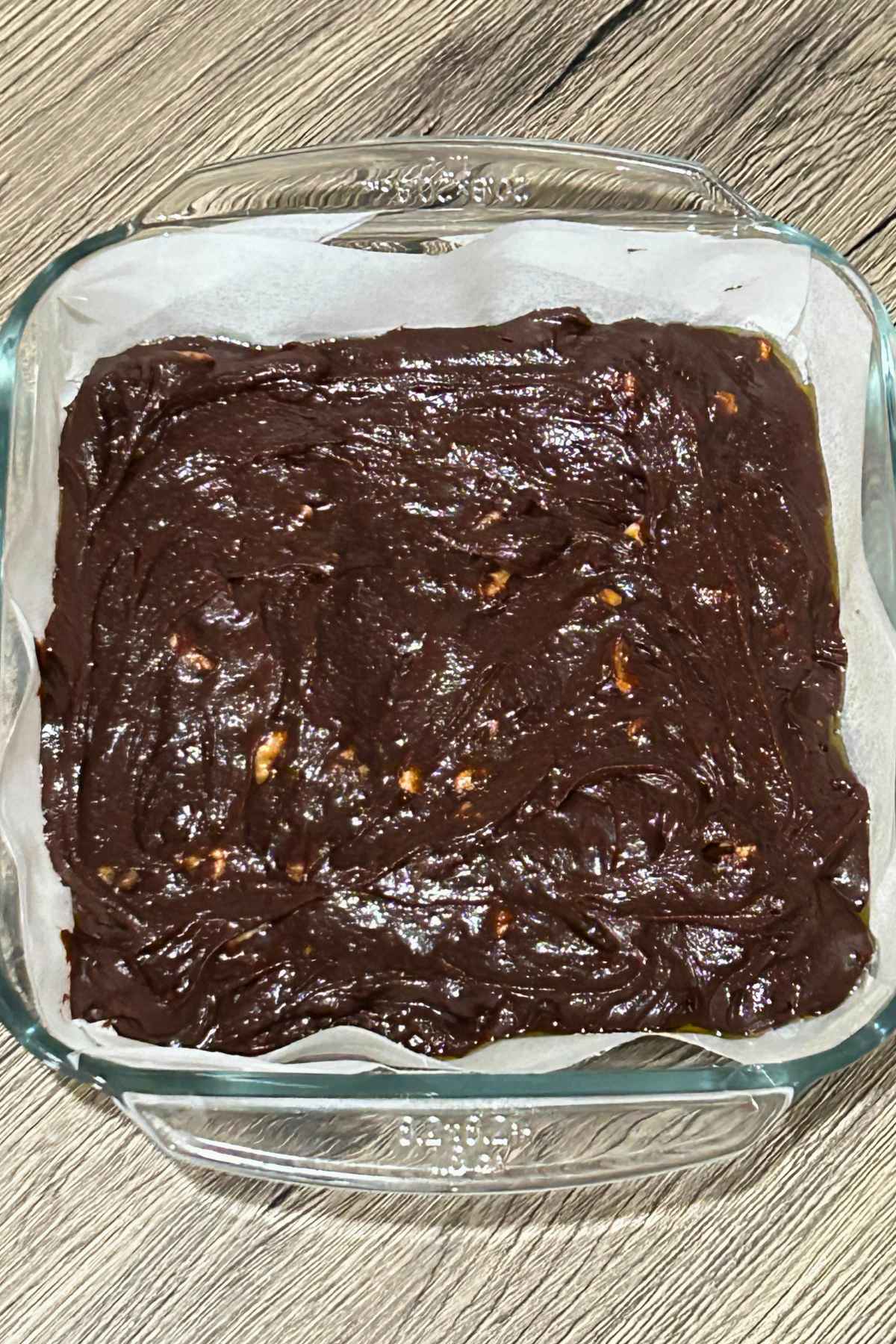 Thick chocolate batter with visible nut pieces spread evenly inside a parchment lined square glass baking dish.