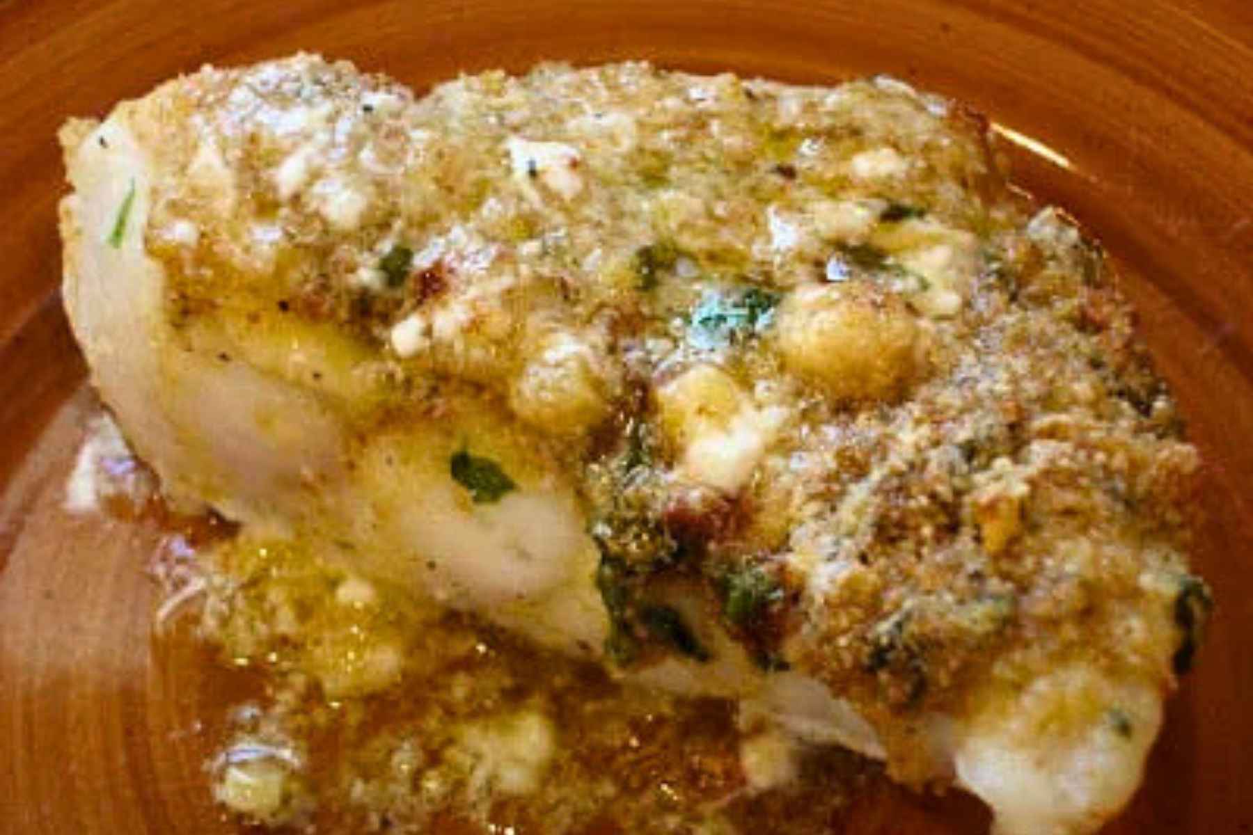 Close up top down fiew of a thick baked red snapper fillet topped with melted butter, herbs, and breadcrumbs on a brown plate.