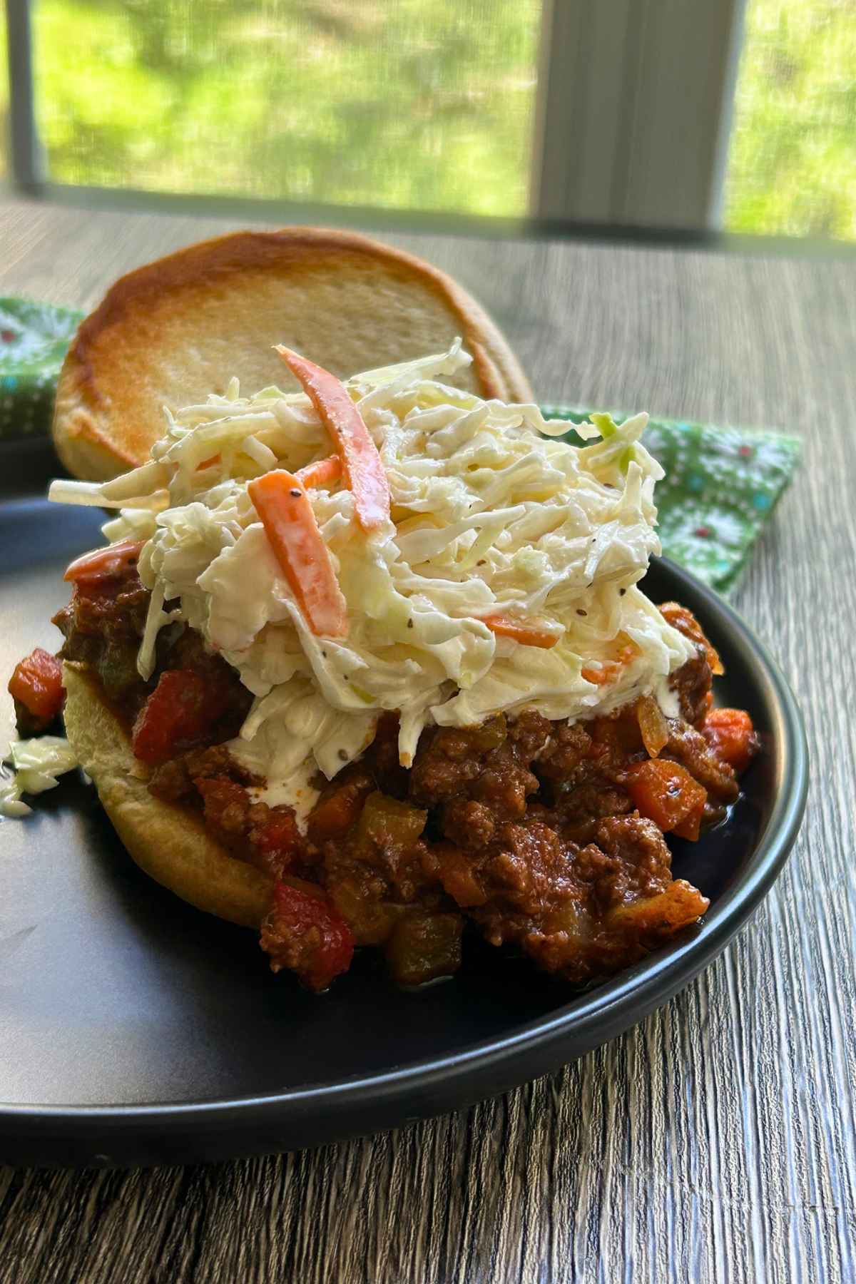A stacked sloppy joe sandwich topped with a generous scoop of creamy coleslaw, served on a black plate with a green patterned napkin in the background.