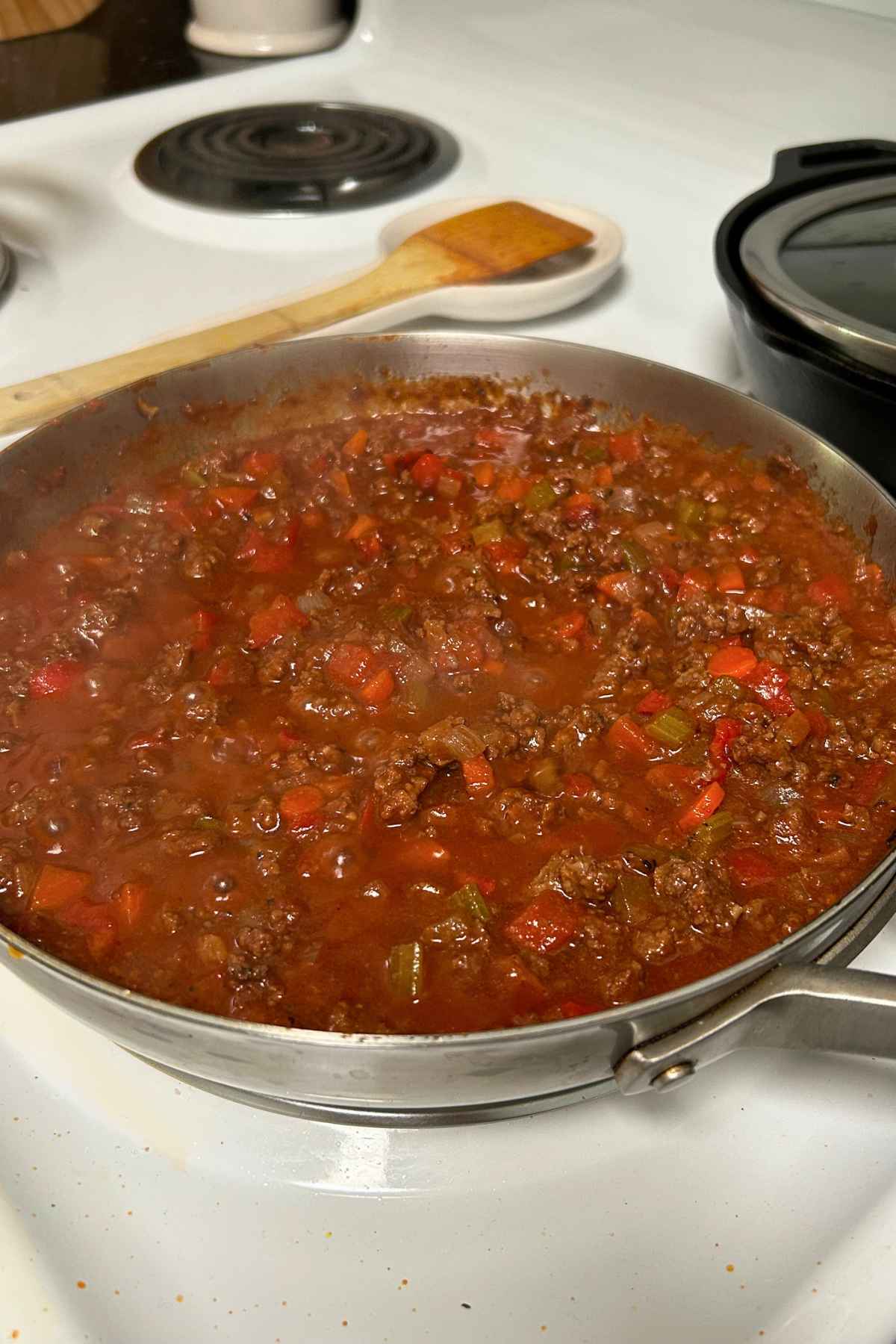 A rich, thick ground beef and chopped vegetable sauce simmering hot in a stainless steel skillet on a white stovetop.