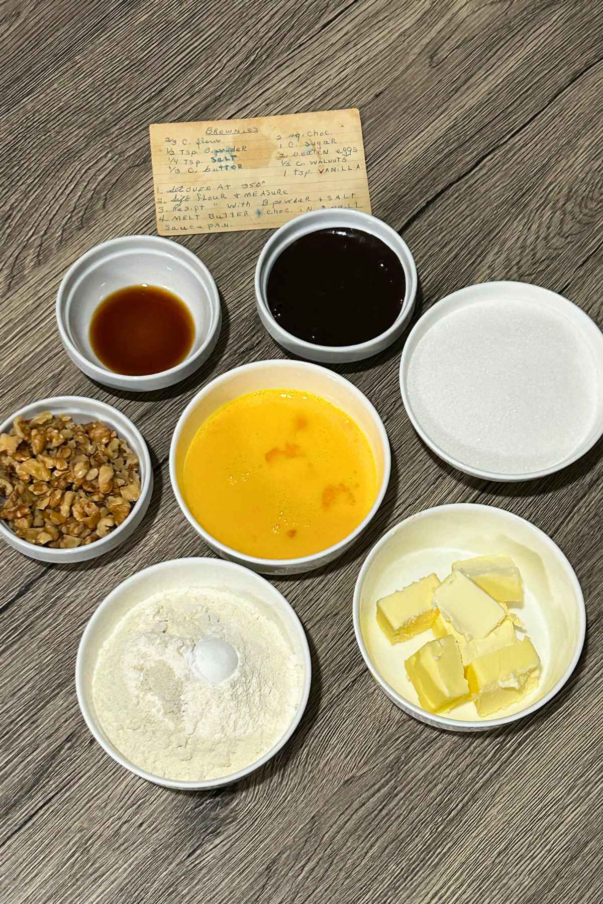 Overhead view of small white prep bowls with flour, butter cubes, egg yolks, chopped walnuts, and liquids arranged around a handwritten recipe card on a dark wooden surface.