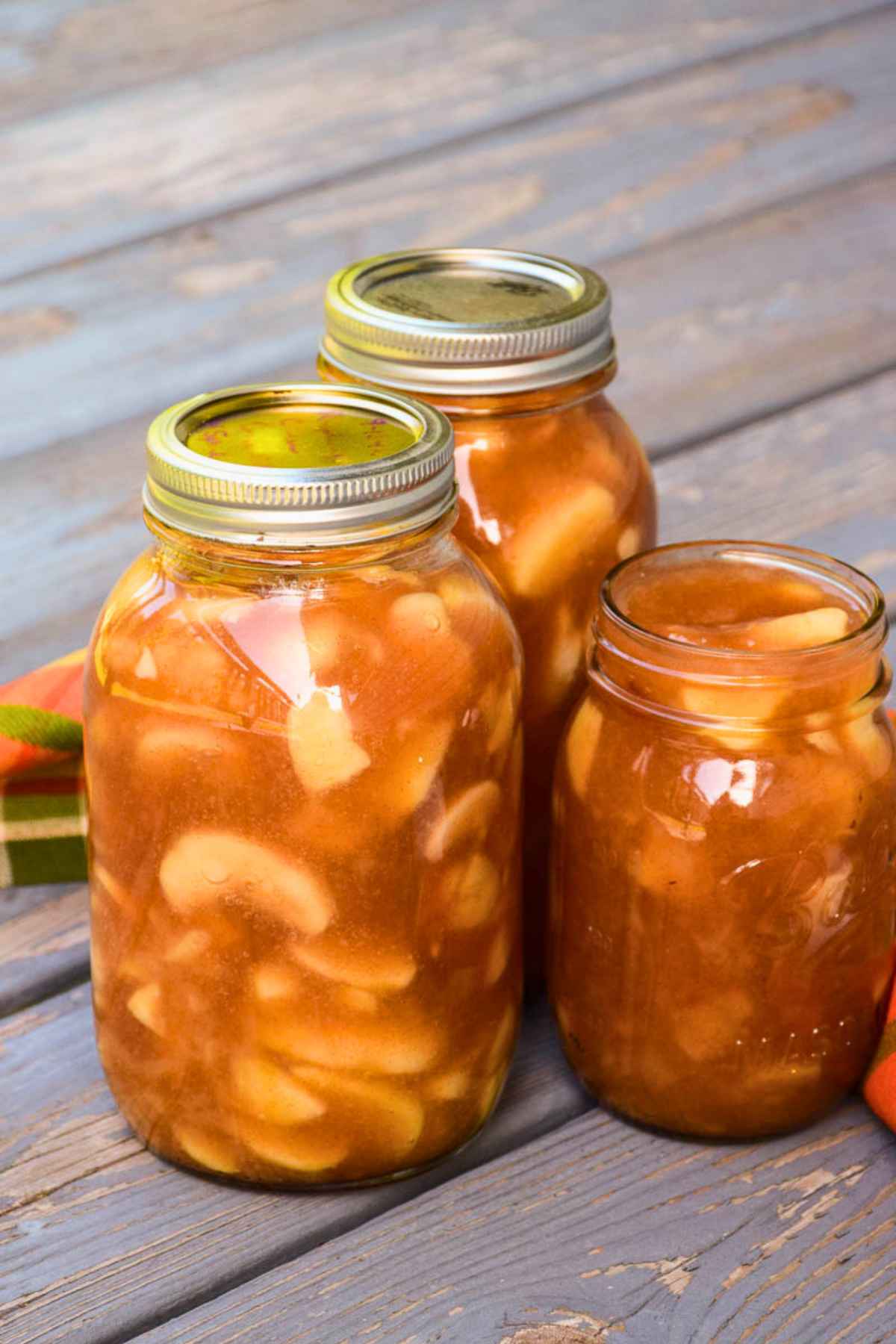 Two quart sized jars and one pint sized jar of homemade apple pie filling on a rustic wood plank with a orange and green plaid napkin in the background.