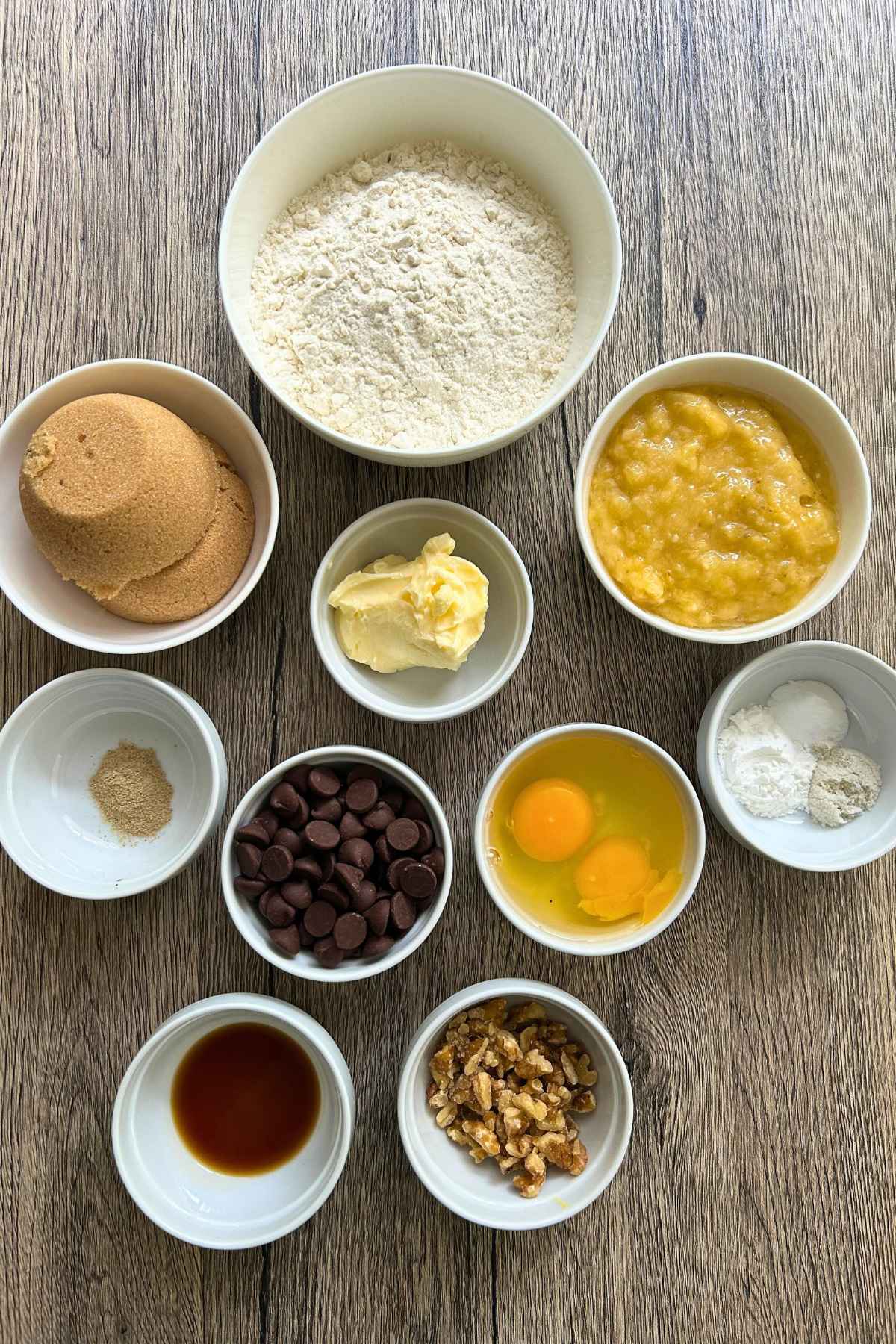 Small white bowls filled with baking ingredients including flour, brown sugar, mashed bananas, butter, eggs, chocolate chips, and spices arranged on a wooden surface.